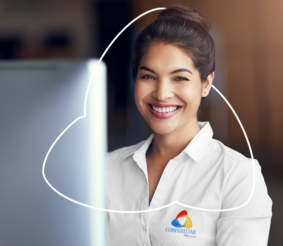Mujer sonriente con camisa blanca y logo de Comfamiliar Atlántico, de pie frente a una computadora en un entorno de oficina iluminado. Fondo desenfocado.