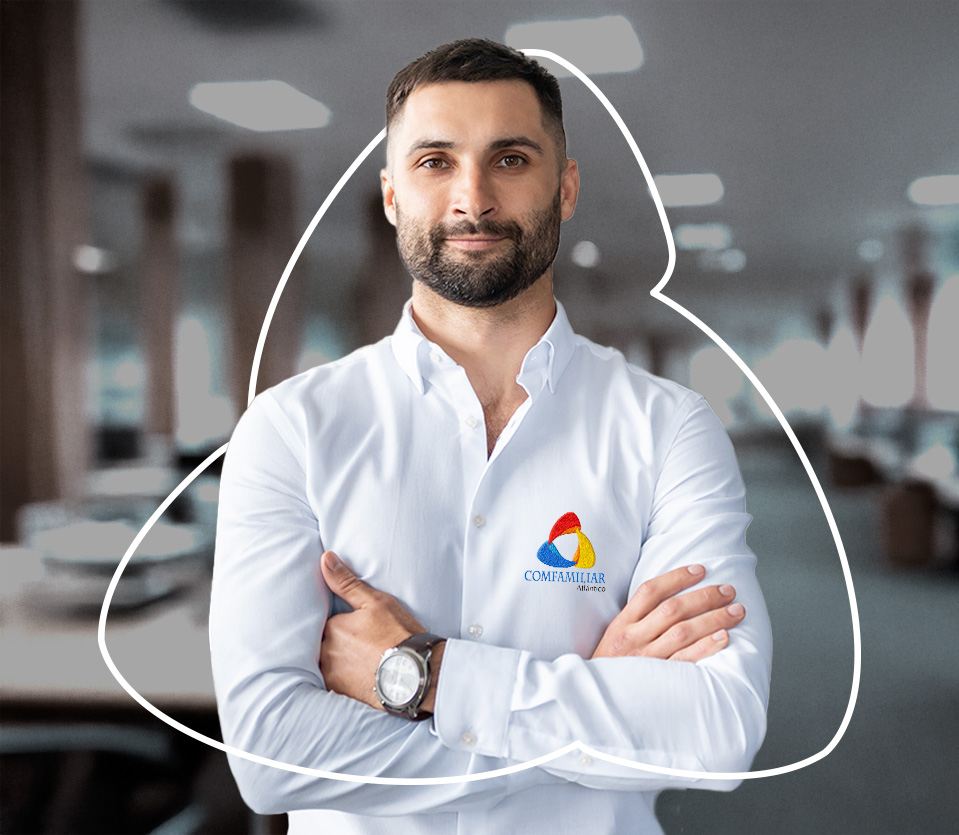 Hombre de pie con camisa blanca y logo de Comfamiliar, brazos cruzados, en un entorno de oficina con mobiliario moderno y luz natural.