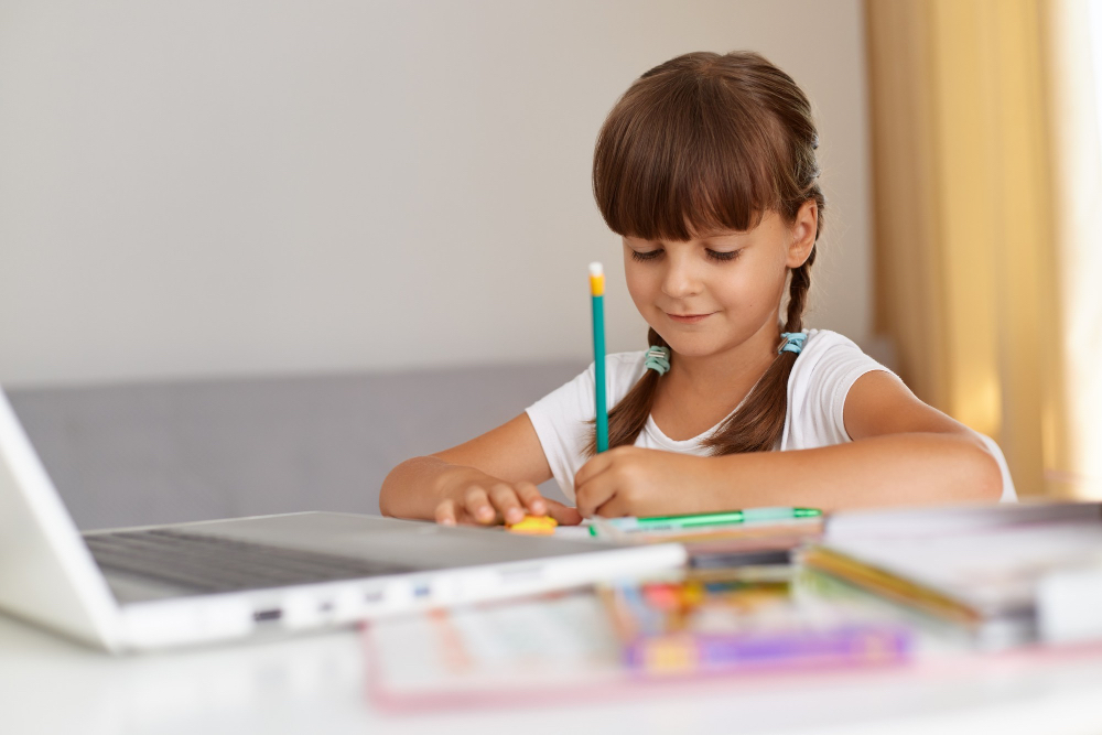 Niña dibujando con lápiz en una mesa, junto a una computadora portátil y materiales de arte coloridos.