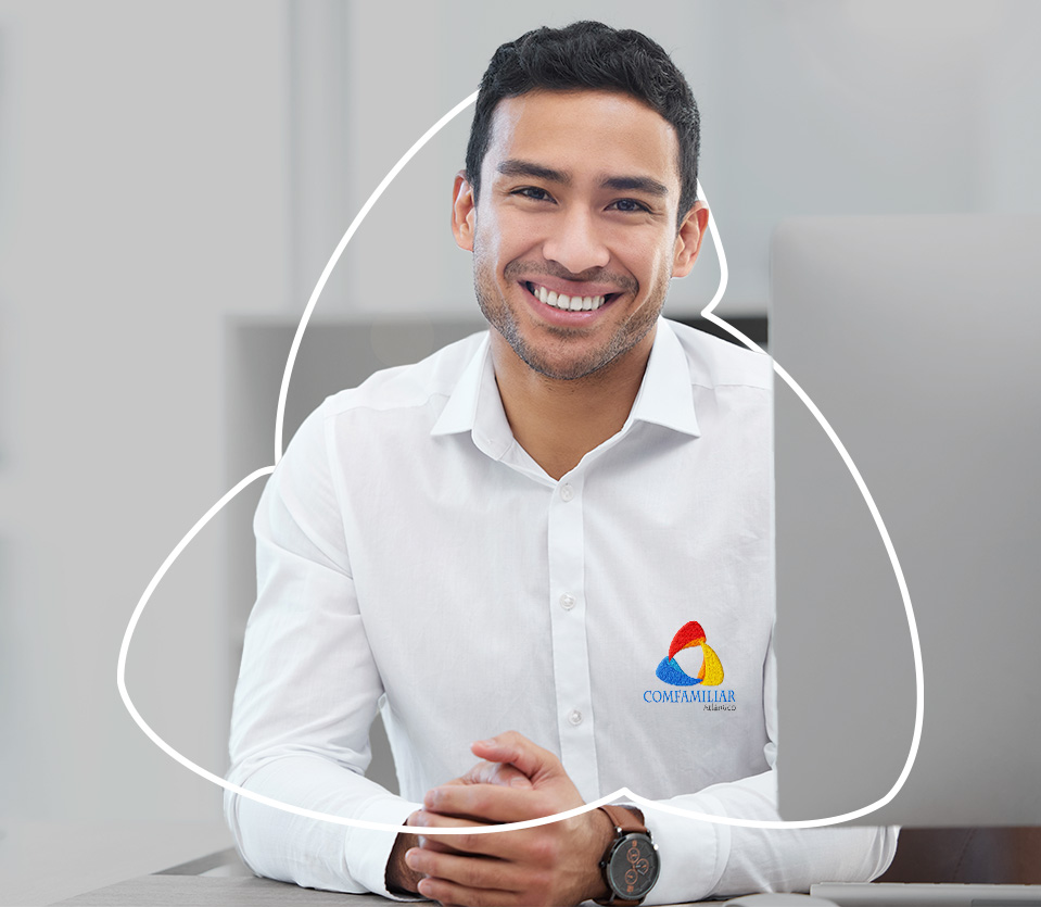 Hombre sonriente con camisa blanca y logo de Comfamiliar, sentado frente a una computadora en un entorno de oficina con tonos neutros.
