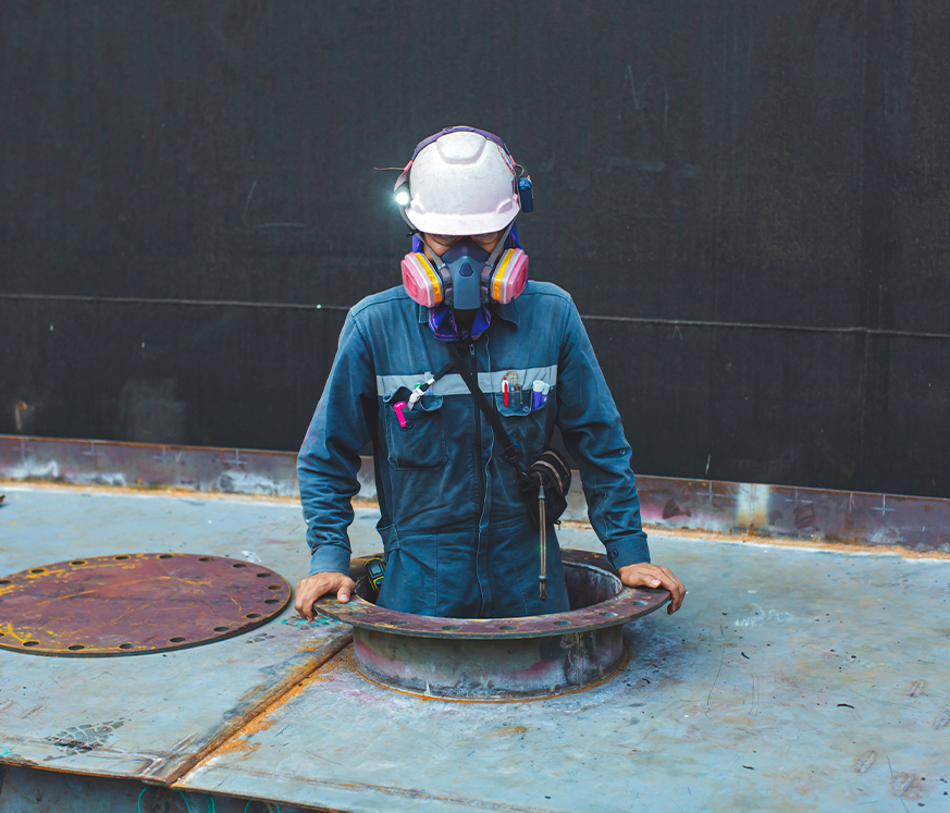 Trabajador con casco y equipo de protección personal, asomándose desde una abertura en una superficie metálica. Fondo oscuro, predominan tonos grises y azules.