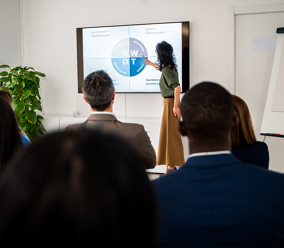 Presentadora señala un gráfico SWOT en pantalla mientras un grupo de personas observa. El entorno es moderno, con una planta verde y un proyector visible. Predominan tonos neutros.