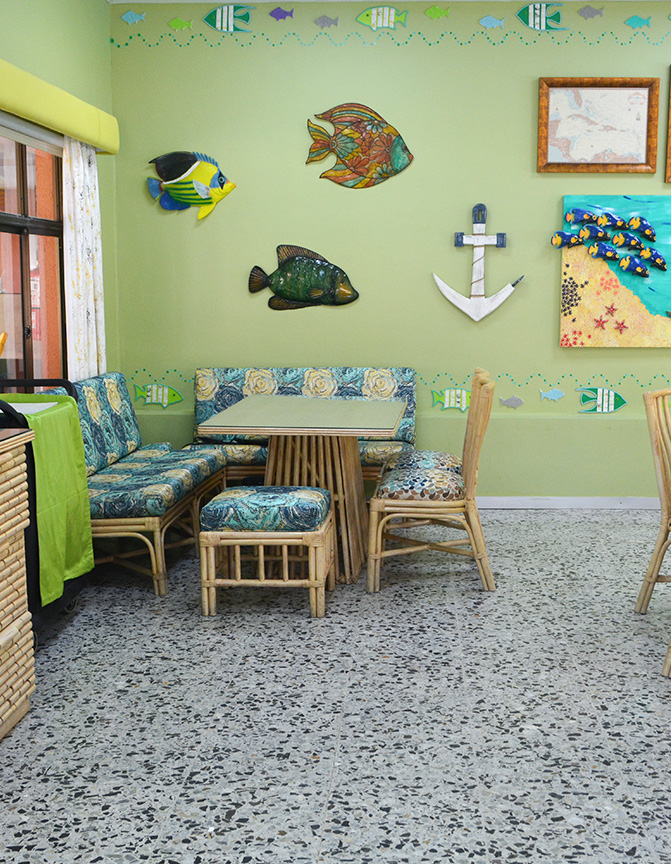 Espacio interior con paredes verdes decoradas con peces y ancla. Mobiliario de madera clara y cojines coloridos. Mesa central rodeada de sillas y un suelo de granito.