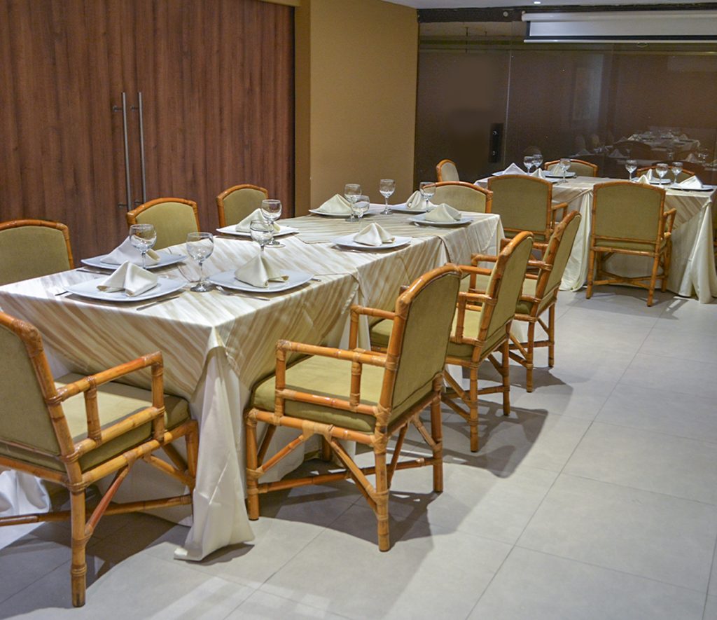 Mesa larga con manteles claros, sillas de madera y cojines verdes, dispuesta para una comida, con copas y platos en cada lugar, en un entorno moderno y bien iluminado.