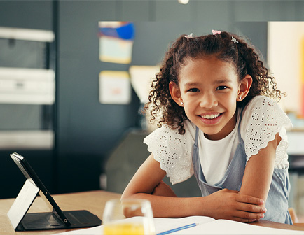 Niña sonriente con cabello rizado, apoyada en una mesa con un dispositivo y un vaso de bebida. Fondo oscuro.