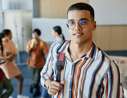 Joven con camisa de rayas y gafas, sonriente, sosteniendo una mochila. En el fondo, otras personas conversan en un entorno educativo con paredes claras y mobiliario moderno.