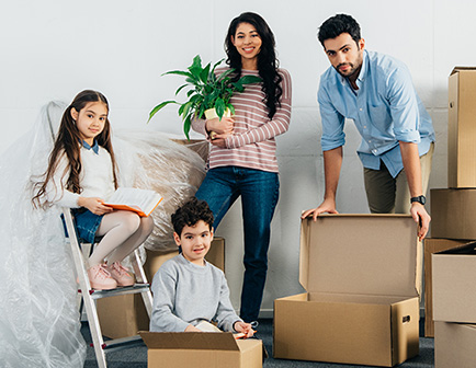 Familia en un entorno de mudanza, con dos adultos y dos niños. Una niña está sentada en una escalera con un libro, mientras los demás sostienen cajas y una planta.