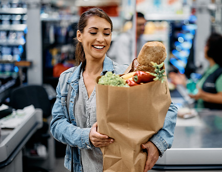 Mujer sonriente sostiene una bolsa de papel con alimentos en un supermercado, con estantes y productos al fondo.