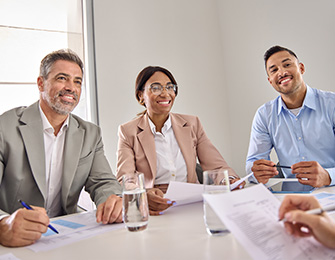 Tres personas sentadas alrededor de una mesa, revisando documentos. Dos hombres y una mujer, todos vestidos formalmente, con botellas de agua y papeles frente a ellos.