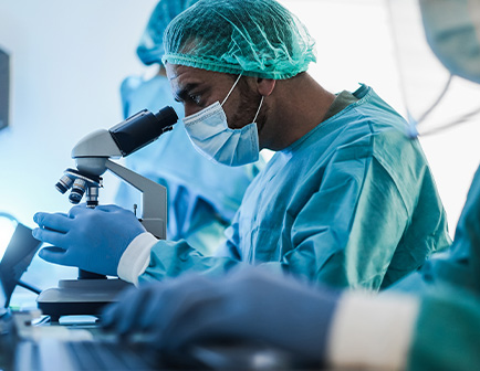 Profesional de la salud en un entorno de laboratorio, usando un microscopio. Viste equipo de protección personal, incluyendo gorro, mascarilla y guantes. Fondo con iluminación suave y equipo técnico.