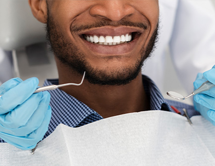 Hombre sonriente en un consultorio dental, con dentista utilizando herramientas de odontología. Se observa un entorno clínico con tonos claros y un enfoque en la salud dental.
