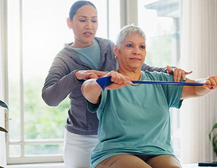 Una mujer mayor realiza ejercicios de rehabilitación con una banda elástica, mientras una instructora la asiste en un entorno luminoso.