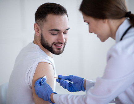 Hombre sonriente recibiendo una inyección en el brazo por parte de una profesional de la salud, en un entorno clínico con tonos claros y luz natural.