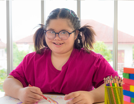 Niña con gafas y coletas, sonriente, dibujando en una mesa con lápices de colores en un entorno iluminado.