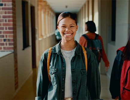 Joven sonriente con mochila en un pasillo escolar, con otras personas al fondo y luz natural.