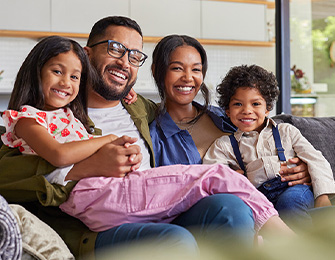 Familia sonriente sentada en un sofá, compuesta por dos adultos y dos niños, en un entorno acogedor y luminoso.