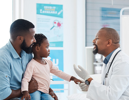 Padre e hija en una consulta médica, el padre sostiene a la niña mientras un médico, con guantes, le habla. Fondo con carteles informativos y colores claros.