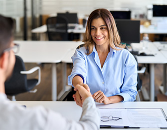 Mujer sonriente con camisa azul estrechando la mano de un hombre, en un entorno de oficina moderno con escritorios y computadoras. Colores neutros predominan en el espacio.