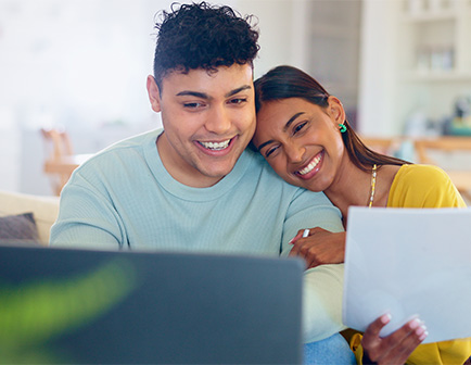 Pareja sonriente usando una computadora portátil en un entorno acogedor, con documentos en mano y colores cálidos predominantes.