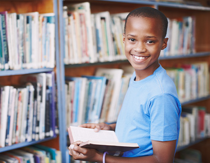 Niño sonriente sosteniendo un libro en una biblioteca, con estantes llenos de libros en el fondo. Lleva una camiseta azul y está en un ambiente iluminado.