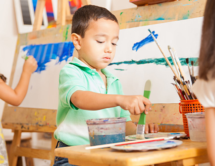 Niño pintando con un pincel verde, rodeado de materiales de arte y otro niño al fondo. Colores brillantes en el entorno.