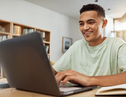 Joven sonriente trabajando en una laptop, con estanterías y un ambiente luminoso al fondo.