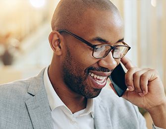 Hombre con gafas, sonriendo mientras habla por teléfono, vestido con traje claro, en un entorno iluminado.