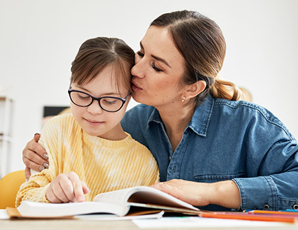 Mujer abraza a una niña que lee un libro, ambas sonríen. La niña lleva gafas y una camiseta amarilla, mientras que la mujer viste una camisa de mezclilla. Fondo claro y acogedor.