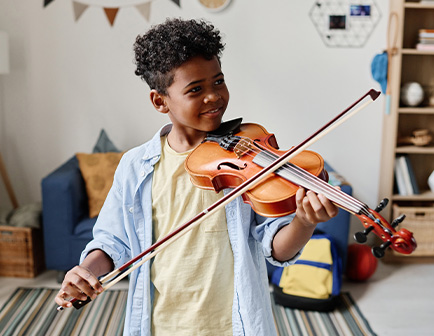Niño tocando un violín, vestido con camiseta amarilla y camisa de mezclilla, en un entorno interior con sofá azul.