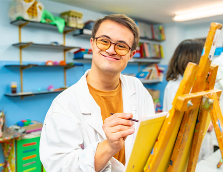 Hombre sonriente con gafas, vestido con bata blanca, sostiene un pincel frente a un caballete en un entorno colorido con materiales de arte y estanterías al fondo.