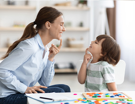 Mujer y niño sonríen mientras juegan, sentados frente a una mesa con materiales coloridos en un ambiente luminoso.