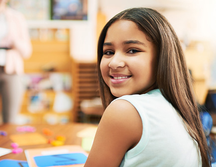Niña sonriendo en un aula, con cabello largo y oscuro, sentada frente a una mesa con materiales escolares. El entorno es luminoso y colorido, con objetos de aprendizaje visibles.