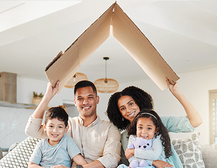 Familia sonriente en un salón, sosteniendo un cartón en forma de techo. Dos niños pequeños y dos adultos están sentados en un sofá, rodeados de un ambiente acogedor y luminoso.