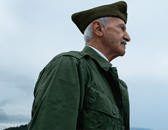 Hombre mayor con gorra y abrigo verde, mirando hacia el horizonte. Fondo con cielo nublado y paisaje difuso.