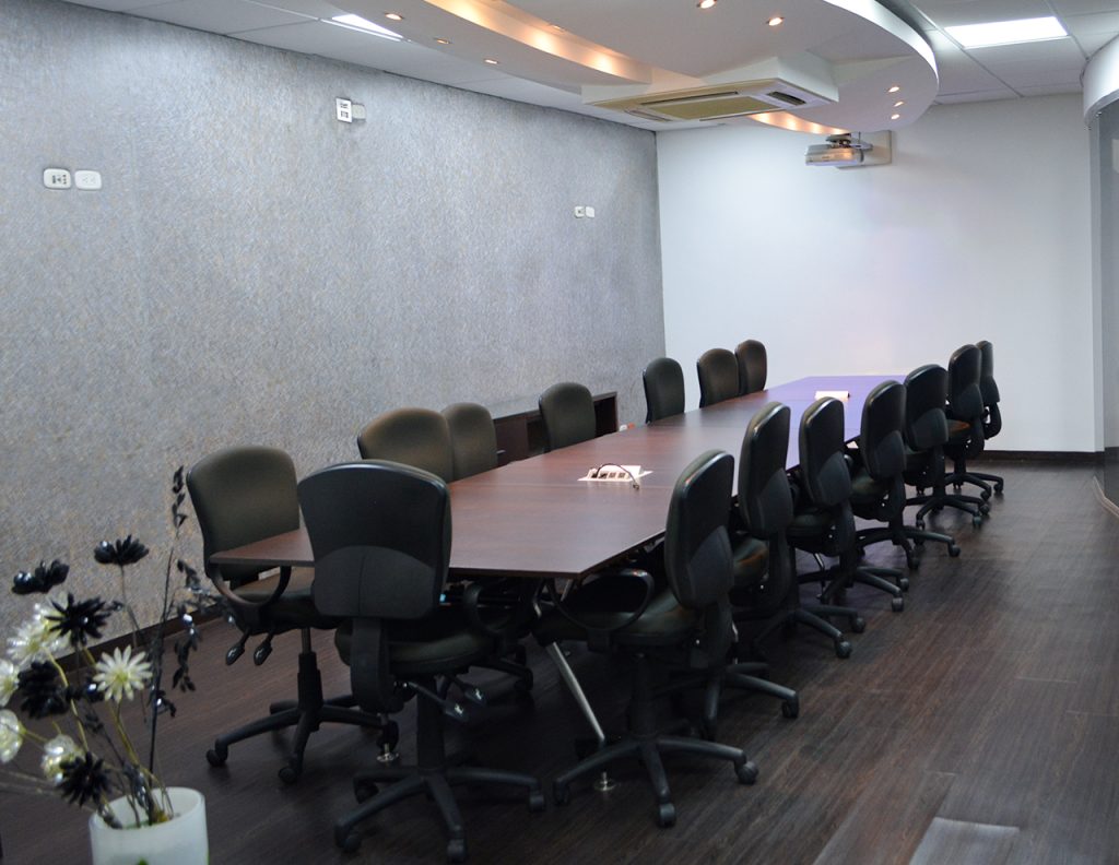 Sala de reuniones con una mesa larga de madera y sillas giratorias negras, decorada con una planta en el extremo. Paredes en tonos claros y suelo de madera oscura.