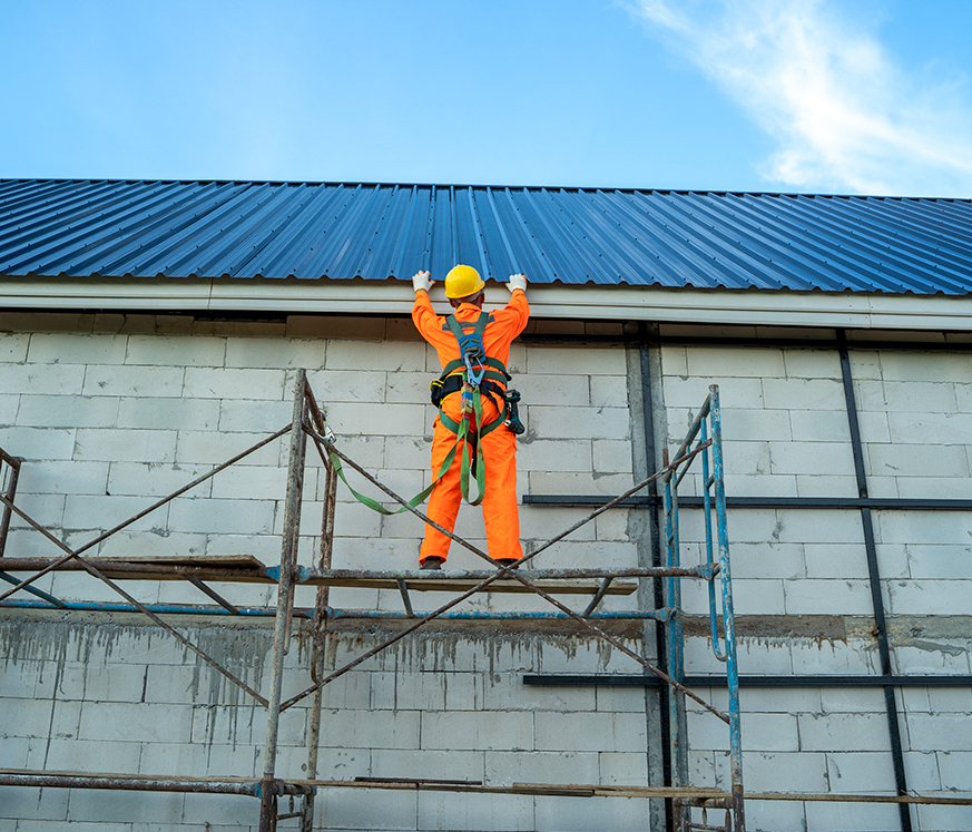 Trabajador en overol naranja y casco amarillo, en andamio, instalando un techo de metal en una pared de bloques grises bajo un cielo azul.