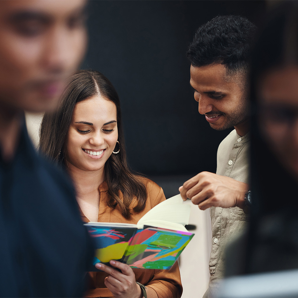 Mujer sonriente sostiene un cuaderno colorido mientras un hombre le muestra una página, en un entorno con otros individuos desenfocados al fondo. Colores cálidos predominan en la vestimenta.