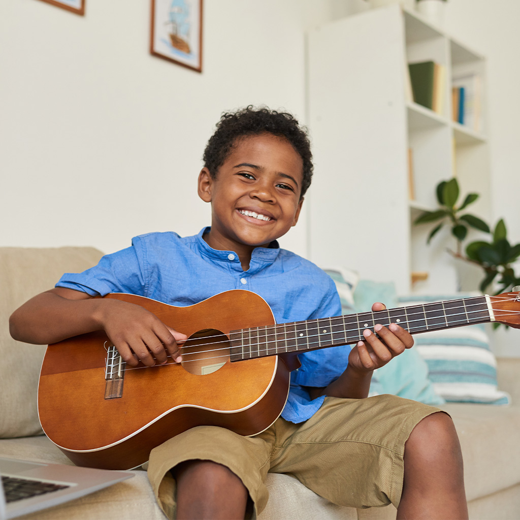 Niño sonriente tocando un ukelele en un sofá, con camisa azul y pantalones cortos. Al fondo, estanterías con libros y plantas, en un ambiente luminoso y acogedor.