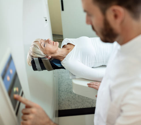 Mujer acostada en una camilla, con un dispositivo en la cabeza, mientras un técnico ajusta un panel de control. El entorno es clínico y luminoso.