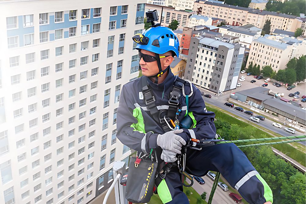 Trabajador en arnés de seguridad y casco, suspendido frente a un edificio alto, observa la ciudad con vehículos y árboles visibles en el fondo. Predominan colores azules y verdes.