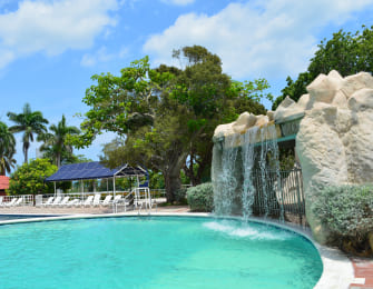Piscina con cascada artificial, rodeada de árboles y áreas de descanso. Cielo parcialmente nublado y tono azul en el agua. Sillas blancas visibles al fondo.