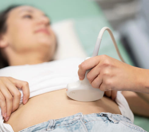Mujer acostada en una camilla, recibiendo un ultrasonido en el abdomen, mientras un profesional sostiene el dispositivo de ultrasonido. Fondo en tonos claros.