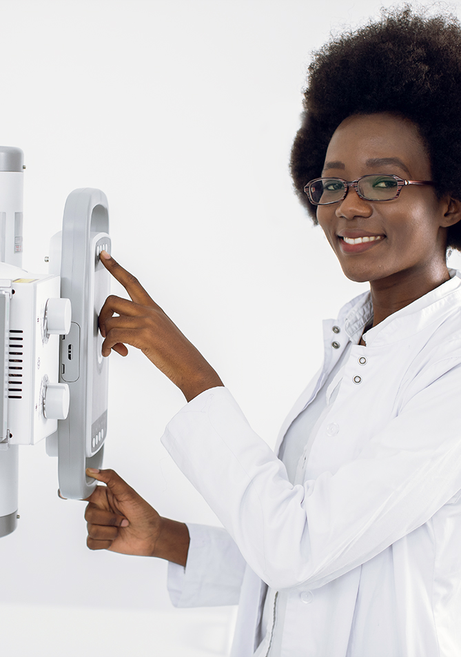 Mujer con cabello rizado y gafas, vestida con bata blanca, interactúa con un equipo médico en un entorno luminoso y minimalista. La máquina muestra botones y pantallas.