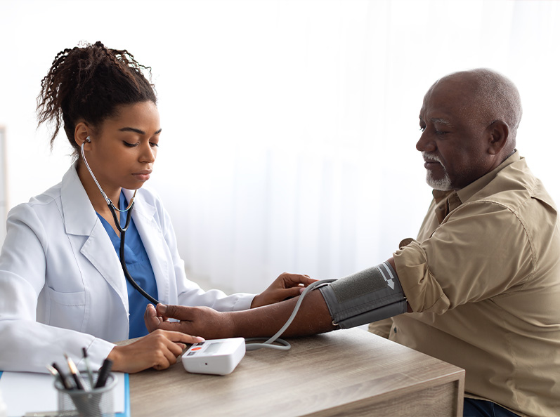 Mujer médica toma la presión arterial a un hombre mayor en un consultorio con luz natural, usando un esfigmomanómetro y un estetoscopio. Ambos están sentados frente a una mesa.