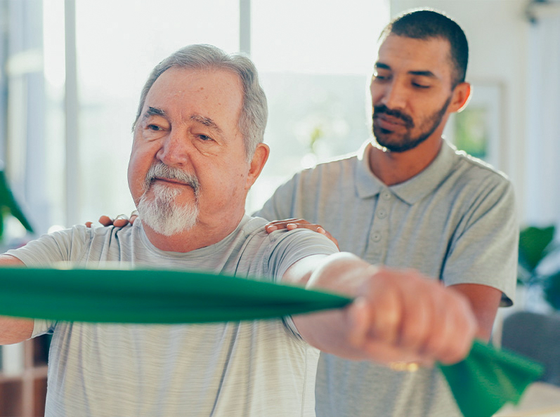 Hombre mayor realizando ejercicios de rehabilitación con una banda elástica, mientras un joven lo asiste desde atrás en un entorno luminoso con plantas decorativas.