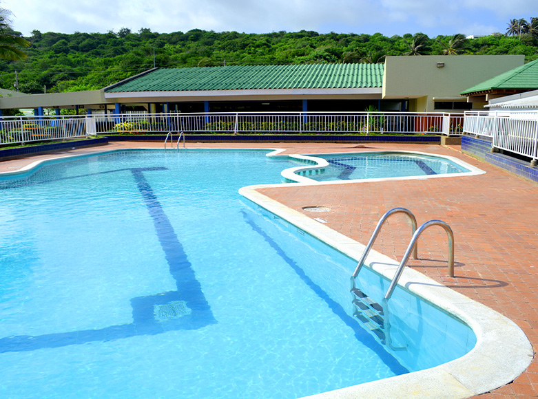 Piscina con escaleras, rodeada de áreas de descanso y vegetación al fondo, bajo un cielo despejado.