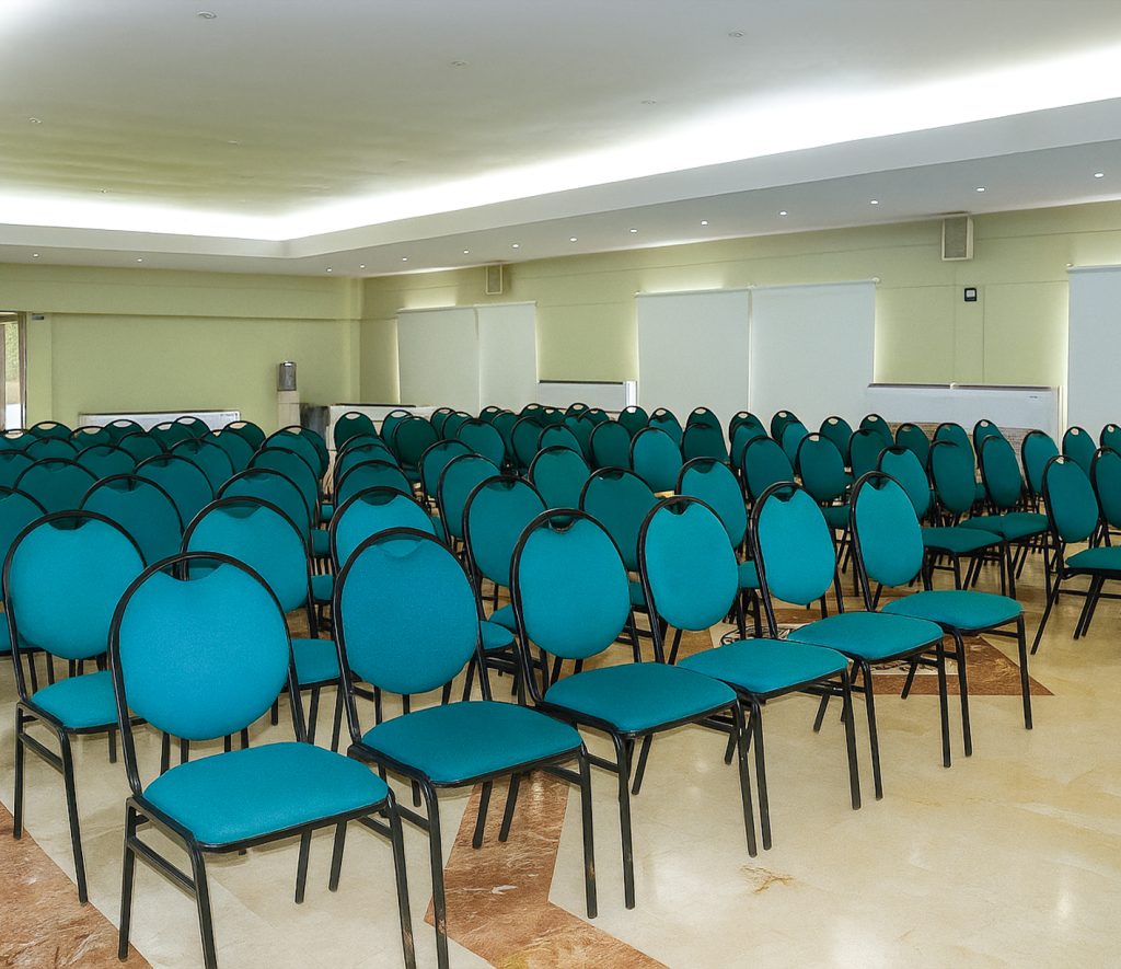 Salón con filas de sillas de color verde azulado, dispuestas ordenadamente sobre un suelo de mármol claro. El ambiente es iluminado y cuenta con paredes en tonos neutros.