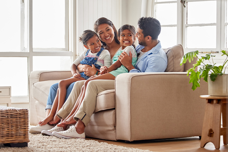 Familia sentada en un sofá, sonriendo y disfrutando juntos. Dos niños pequeños en brazos de sus padres. Ambiente luminoso con ventanas y una planta en la esquina. Colores suaves y cálidos.