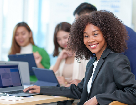 Mujer con cabello rizado sonriente frente a una computadora portátil, mientras tres personas más discuten en un entorno de oficina con colores neutros y elementos tecnológicos.