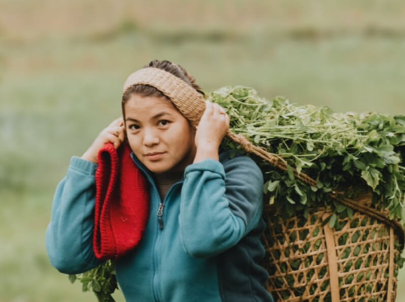 Joven con cabello recogido, viste chaqueta azul y lleva una cesta de mimbre cargada de vegetación, sosteniendo un abrigo rojo mientras mira hacia la cámara en un entorno natural.